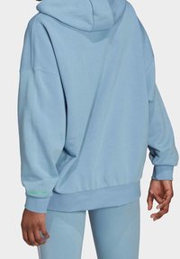 Ljusblå oversized hoodie i mjukt material, med dragsko i huvan, ribbstickade muddar och en grön Adidas-logga på ärmen.