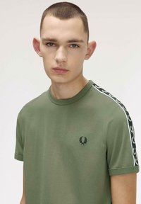Jeune homme aux cheveux courts portant un t-shirt vert olive avec un logo et une bande noire ornée de motifs de laurier blancs sur les manches, fond neutre.