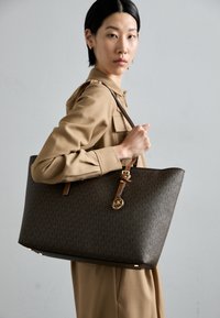 Sac fourre-tout marron à motifs avec des accents en cuir, affichant un pendentif logo doré et une forme structurée. Le modèle le porte avec une blouse de couleur neutre.