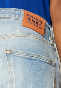 Jasnoniebieskie dżinsy z denimu z naszywką ze skóry z napisem "SCOTCH & SODA AMSTERDAM." Charakteryzują się żółtym przeszyciem oraz klasycznym fasonem z pięcioma kieszeniami.