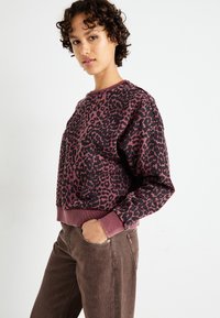 Leopárdmintás pulóver sötét rózsaszín és fekete színben, bordázott mandzsettákkal és aljjal. Barna farmerral viselve. Egyszerű, hétköznapi dizájn.