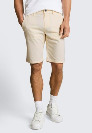 Man die beige knielange shorts, witte sportsokken, witte sneakers en een wit overhemd draagt, met zijn handen in zijn zakken.