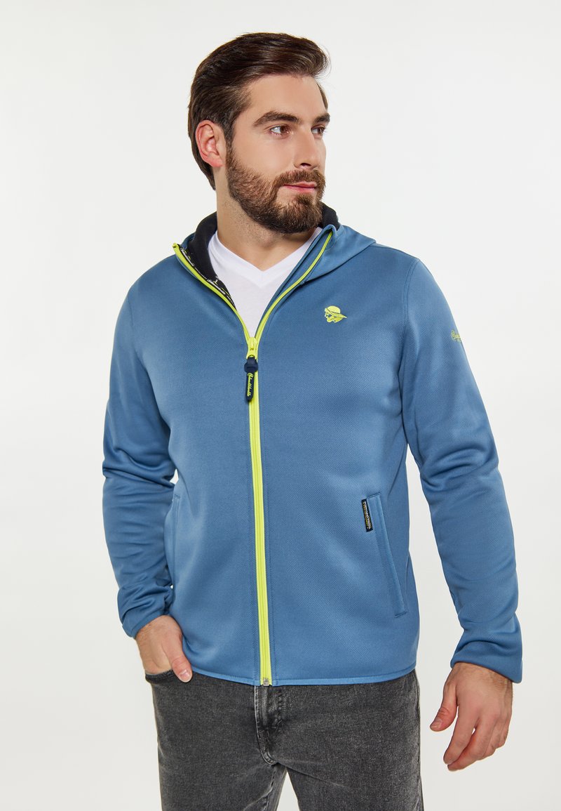 Felpa blu con zip, realizzata in tessuto liscio. Presenta un cappuccio foderato in nero, una zip gialla brillante e un logo sul petto. Accompagnata da una maglietta bianca.