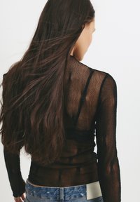 Top negru transparent cu maneci lungi, dintr-un material texturat, având un design ajustat și curele subtiri vizibile pe spate, purtat cu blugi.