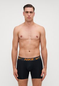 Melnas Nike bokseru bikses ar oranžu logotipu uz jostas. Audums ir gluds un pieguļošs, ar vidēji augstu garumu līdz augšstilbiem.