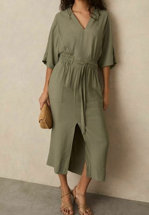 Robe vert olive avec un décolleté en V, une taille élastique et une fente sur le côté. Le matériau a une texture douce. Portée avec des sandales beiges à lanières et un sac en osier.