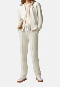 Witte rits-hoodie met een capuchon, gecombineerd met bijpassende slim-fit broek. Gestreept beige en witte shirt eronder. Witte sneakers met bruine zolen.