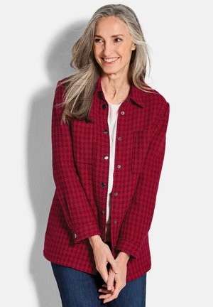 Rood geruiten overhemdjack met een gestructureerde stof, twee borstzakken en zwarte knopen. Draagt boven een witte top en gecombineerd met blauwe jeans.