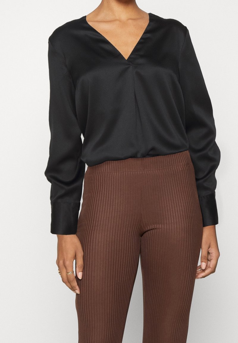 Blouse en satin noir à col en V, manches longues, avec un pli discret à l'avant, associée à un pantalon marron côtelé à taille haute.
