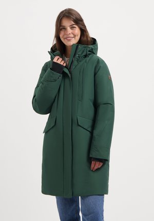 Vrouw glimlachend in een groene gewatteerde winterjas met klepzakken, zwarte manchetten en blauwe jeans tegen een effen witte achtergrond.
