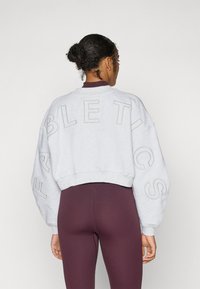 Lys grå kort sweatshirt med oversized passform, med "FABLETICS" trykket på baksiden i stor, uthevet skrift. Kombinert med burgunder tights.