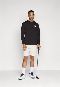 Duga crna majica s dugim rukavima s logotipom, bijele kratke hlače i bijele tenisice s plavim detaljima. Osoba drži golf štap i nosi bijele čarape.