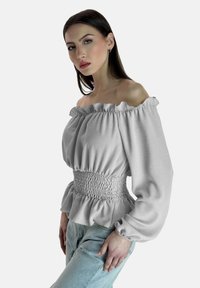 Blusa grigia chiara off-the-shoulder con collo arricciato e vita smock. Maniche lunghe e voluminose con polsini elasticizzati. Indossata sopra a jeans blu.