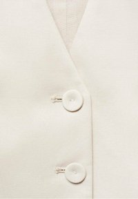 Vêtement beige sans manches avec un col en V, comportant deux boutons ronds et un tissu texturé. Design simple avec peu de détails.