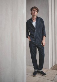 Jonge man staand tussen betonnen pilaren, gekleed in een donker gestreept shirt, wit ondershirt, donkere spijkerbroek en marineblauwe instapschoenen.