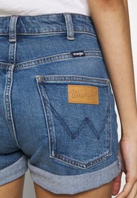 Blå denimshorts med uppvikt kant, med en brun läderlogotyp, bakficka med zigzag-stickning och en Wrangler-tag på midjebandet.