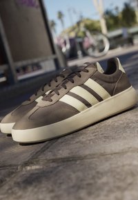 Bruine en beige Adidas sneakers met crèmekleurige zolen op een stenen bestrating, met een wazige stedelijke achtergrond en een fiets.