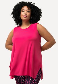 Mouwloze, loszittende top in felroze, gemaakt van een gladde stof. Zijsplitten verhogen het comfort. Gecombineerd met zwarte broek met roze stippen.