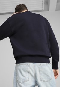 Sudadera azul marino con escote de barco, dobladillo acanalado y puños. Llevada sobre unos jeans de mezclilla azul claro, mostrando un ajuste relajado y textura.