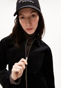 Sort fløjljakke med en sølv lynlås og lomme, lagd ovenpå en mørkebrun turtleneck. Sort kasket med hvidt broderet tekst.