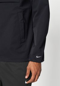 Lähikuva henkilöstä, joka pukeutunut mustaan Nike-takkiin ja -housuihin. Vasemman käden ja käsivarren alue näkyvissä, valkoiset Nike-logot hihan ja housun lahkeen kohdalla.