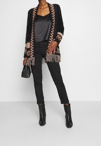 Musta neulecardigan, jossa on geometrisia kuvioita ja reunoja; yhdistetty tumman satiiniseen puseroon ja mustiin kapeneviin housuihin, viimeistelty mustilla nilkkureilla.