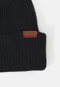 Berretto lavorato a maglia nero con texture a coste, dotato di un'etichetta in pelle marrone impresso con la scritta "Columbia" sul lato.