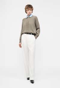 Γυναίκα με μπεζ πουλόβερ πάνω από μπλε πουκάμισο με γιακά, λευκό, ψηλόμεσο παντελόνι με φαρδιά πόδια, μαύρη ζώνη και μαύρα μυτερά παπούτσια, όρθια με τα χέρια στις τσέπες.