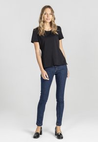 Blondynka stojąca w czarnym topie z krótkim rękawem, ciemnoniebieskich slim fit jeansach i czarnych loafersach na obcasie na jednolitym jasnym tle.