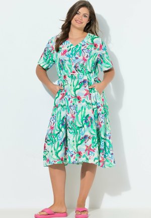 Mujer con un vestido de manga corta y longitud media con estampados coloridos de plantas submarinas y criaturas marinas, con las manos en los bolsillos, sonriendo, de pie.