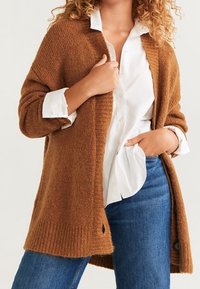 Lang, brun strikket cardigan med ribbede manchetter, åben front og rund nederste kant. Bæres over en hvid skjorte og blå jeans, med subtil tekstur.