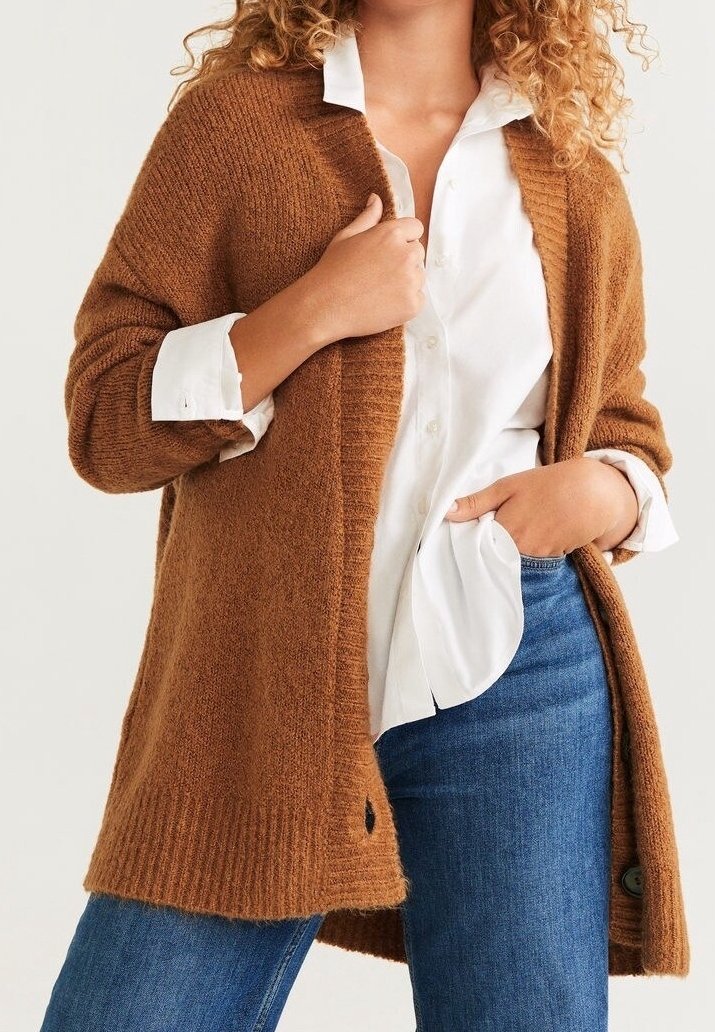 Lang, brun strikket cardigan med ribbede manchetter, åben front og rund nederste kant. Bæres over en hvid skjorte og blå jeans, med subtil tekstur.