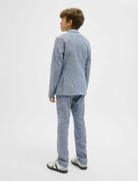 Világoskék, textúrázott öltöny, kifinomult, slim-fit kialakítással; tartalmaz egy zakót és hozzá illő nadrágot. Fehér és fekete sportcipővel viselve.