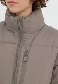 Chaqueta de plumón gris claro con un alto cuello, cierre frontal de cremallera con tirador texturizado y un patrón acolchado para mayor aislamiento.