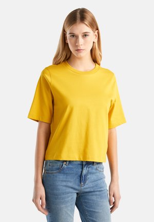 Młoda kobieta o prostych blond włosach, ubrana w żółtą koszulkę z krótkim rękawem i niebieskie dżinsy, stojąca na jasnym tle.