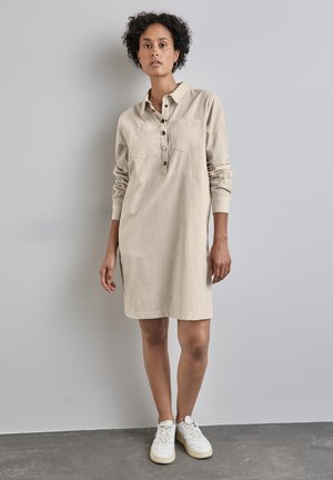 Robe chemise beige à manches longues avec col, fermeture à boutons et deux poches poitrines. Portée avec des baskets blanches sur un fond gris.
