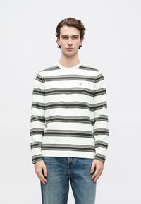 Νεαρός άνδρας κοιτάζει μπροστά, φορώντας λευκό μακρυμάνικο πουκάμισο με μαύρες οριζόντιες ρίγες και μπλε τζιν, σε έναν απλό ανοιχτό φόντο.