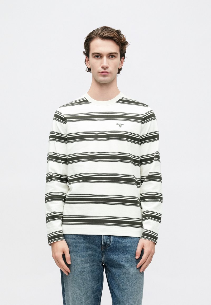 Νεαρός άνδρας κοιτάζει μπροστά, φορώντας λευκό μακρυμάνικο πουκάμισο με μαύρες οριζόντιες ρίγες και μπλε τζιν, σε έναν απλό ανοιχτό φόντο.