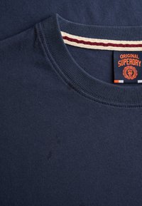 Granatowa bluza z okrągłym dekoltem, prążkowanym kołnierzem i pomarańczową metką "Original Superdry" na wewnętrznej tylnej stronie kołnierza.