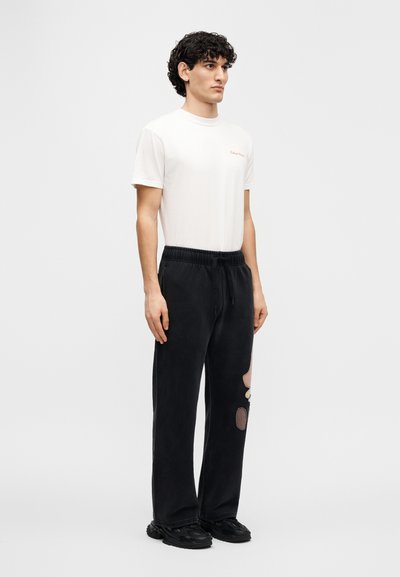 Μαύρο παντελόνι φόρμας με μεγάλα μπαλώματα, ελαστική μέση, συνδυασμένο με εφαρμοστή λευκή μπλ ούζα που φέρει κόκκινο κείμενο στο στήθος.