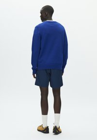 Pull en tricot bleu avec poignets côtelés, associé à un short bleu marine. Chaussures noires avec des accents jaunes et chaussettes blanches visibles. Fond minimaliste.