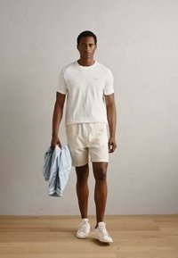 Witte katoenen t-shirt, beige shorts en witte sneakers. Houdt een lichtblauw gestreept shirt vast. Staat op houten vloeren tegen een neutrale muur.