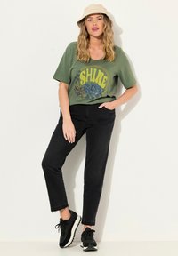 T-shirt verde oliva con stampa gialla che recita "SHINE," abbinata a jeans neri e sneakers neri, completata da un cappello a secchiello beige chiaro.
