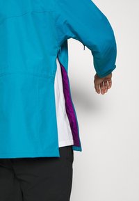 Personne portant une veste bleu sarcelle dézippée sur le côté, révélant une doublure intérieure violette et une chemise blanche en dessous.