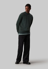 Sudadera verde scuro con polsini e orlo a coste, sovrapposta a un tessuto più chiaro. Abbinata a pantaloni neri larghi e scarpe nere.