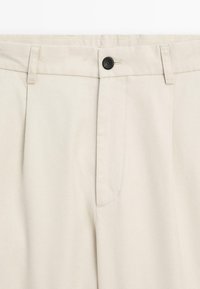 Beige trousers with a smooth texture, featuring a single black button, two front pleats, and side pockets for functionality.