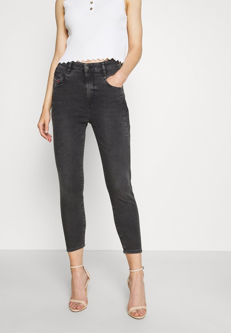 Jeans negros de talle alto, ajuste slim, con bolsillos frontales y costuras rojas sutiles, combinados con un top blanco corto y sandalias nude de tacón alto.