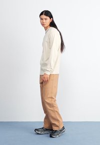 Lys beige langermet skjorte, brune bukser og svarte joggesko; modellen står sidelengs mot en hvit vegg på et blått gulv.