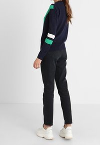 Granatowy sweter z dzianiny z zielonymi i białymi paskami na rękawach, w połączeniu z czarnymi spodniami i białymi trampkami, widziany od tyłu.