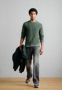 Grüner, gerippter Pullover, dunkelgraue Jeans, hält eine grüne Bomberjacke; leichte Sneakers mit gemusterten Akzenten, Holzfußboden im Hintergrund.